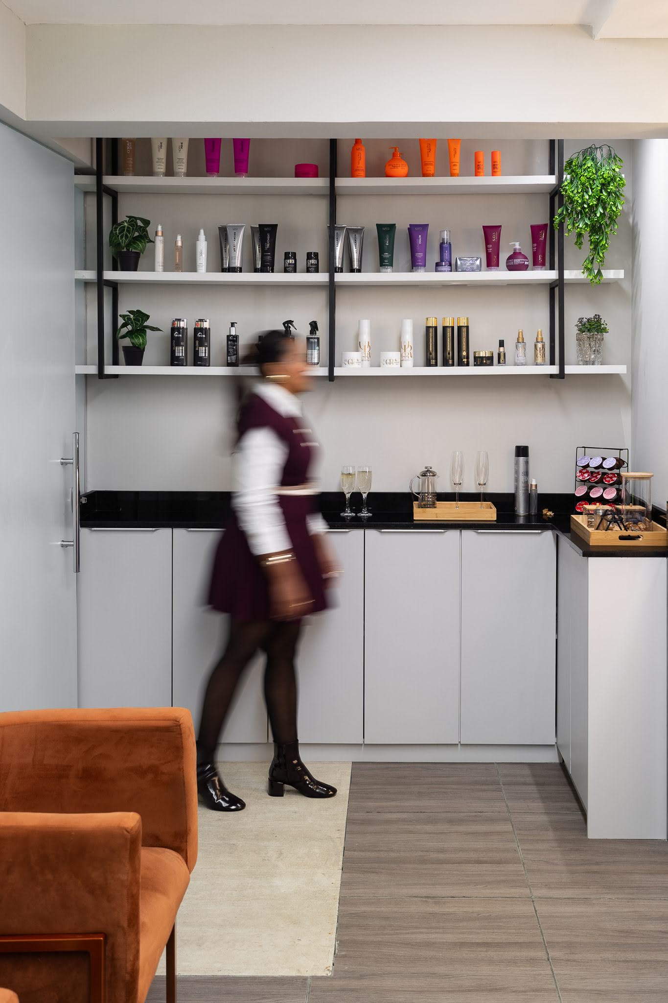 Estante com cosméticos e cafeteira sobre bancada preta, com cliente ao fundo